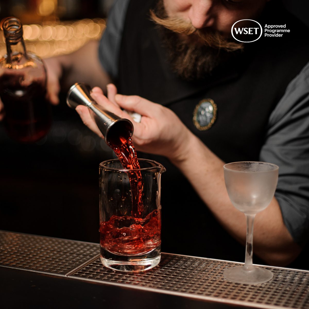 Bartender pouring amber spirit from a jigger into a mixing glass at a bar