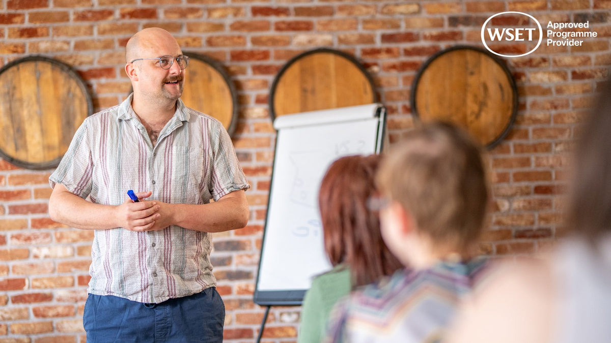 WSET educator presenting beside a flipchart in a spirits classroom with wooden barrel ends on a brick wall
